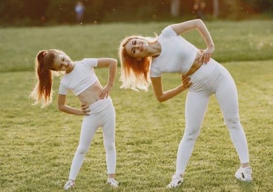 A mother and daughter in activewear stretching on a sunny day in a grassy field.