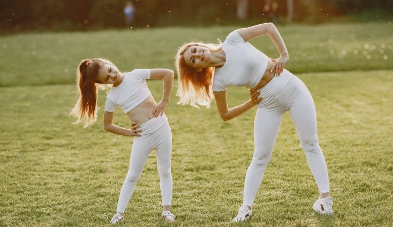 A mother and daughter in activewear stretching on a sunny day in a grassy field.